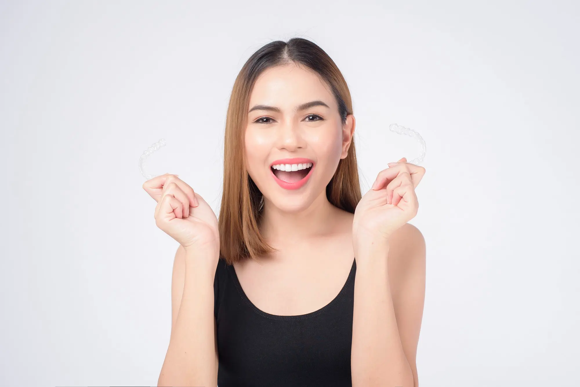 Excited young woman holding clear Invisalign aligners in both hands, showcasing the transparent, removable orthodontic treatment option available at Sweet Smiles Orthodontics in Arlington, MA for discreet teeth straightening
