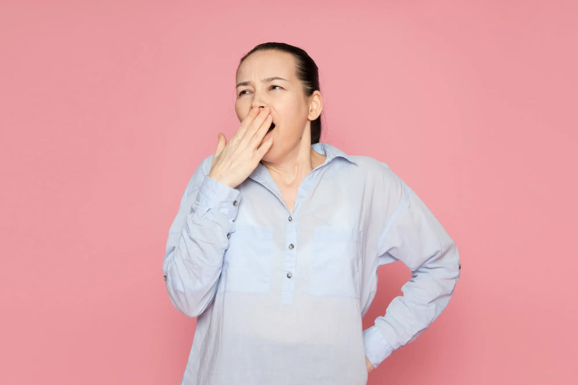 Against a pink background, woman in blue shirt yawns with hand over mouth breathing in Arlington, MA.