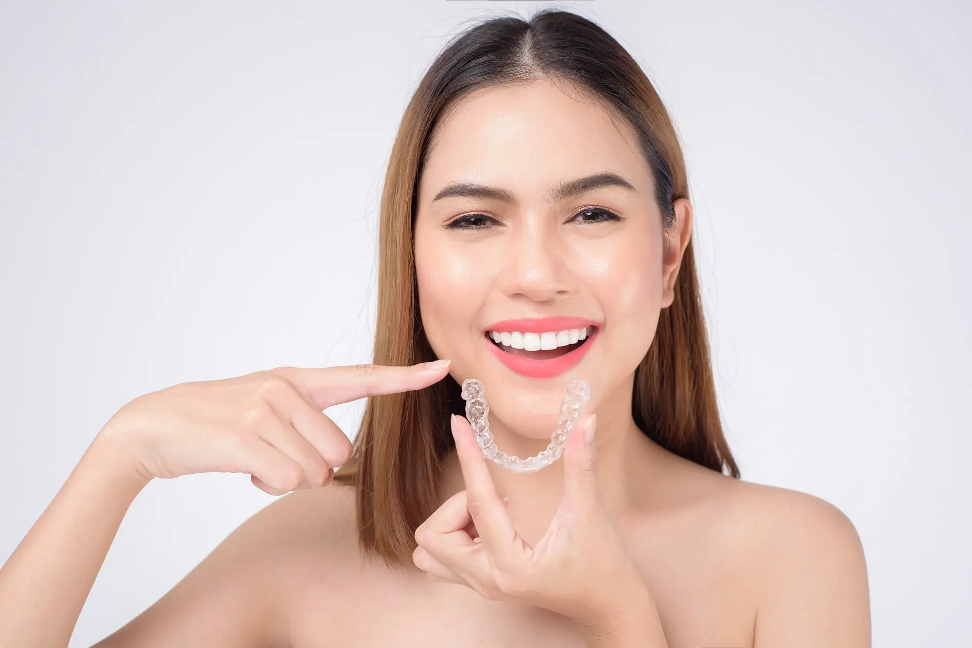 Smiling young woman pointing to her beautifully aligned teeth while holding her Invisalign clear aligner, showcasing successful orthodontic treatment made affordable through Invisalign Insurance coverage at Sweet Smiles Orthodontics in Arlington, MA