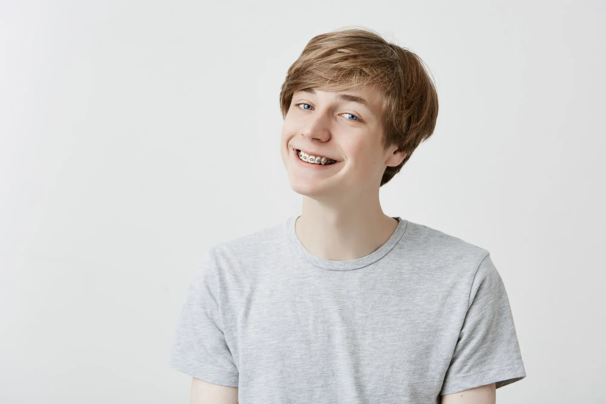 A smiling teen with light brown hair and braces in a gray t-shirt, photographed at Sweet Smiles Orthodontics in Arlington, MA.