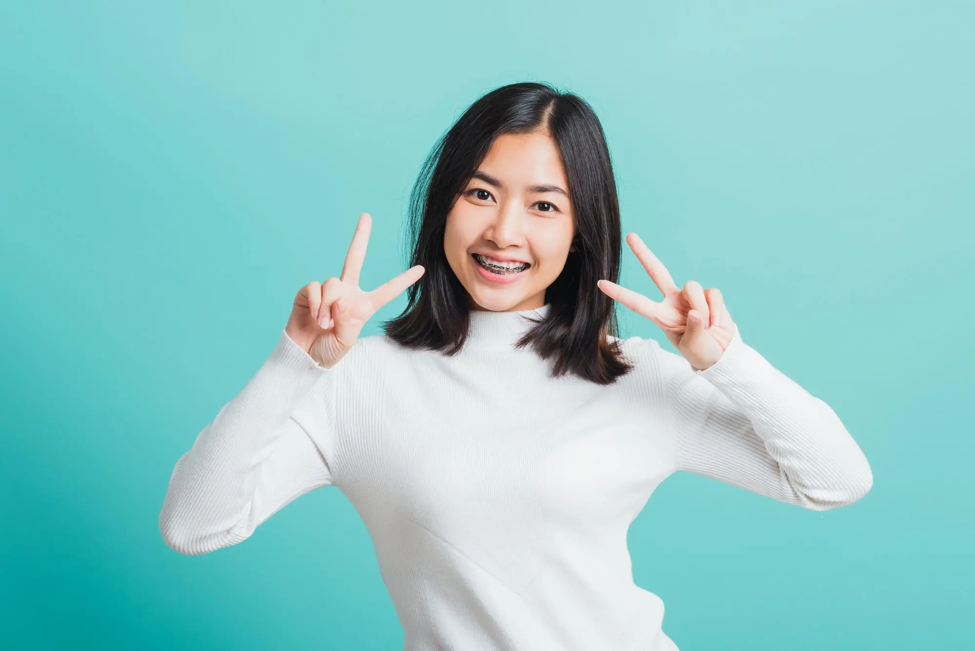 At Sweet Smiles Orthodontics in Arlington, MA, a young woman with metal braces smiles and shows peace signs against a light blue background.