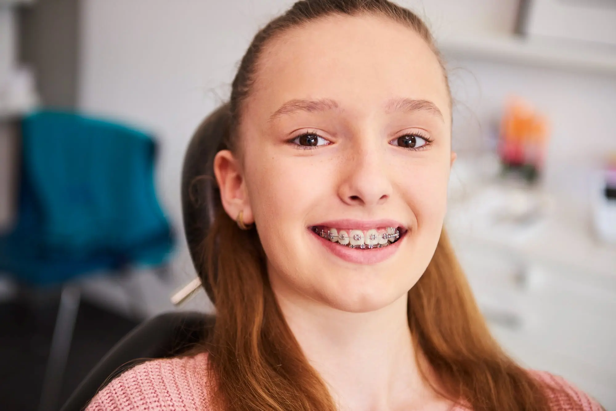Sweet Smiles Orthodontics Arlington - Braces & Invisalign Smiling girl with long brown hair and affordable braces cost sits in a Sweet Smiles Orthodontics chair in Arlington, MA; dental equipment behind.