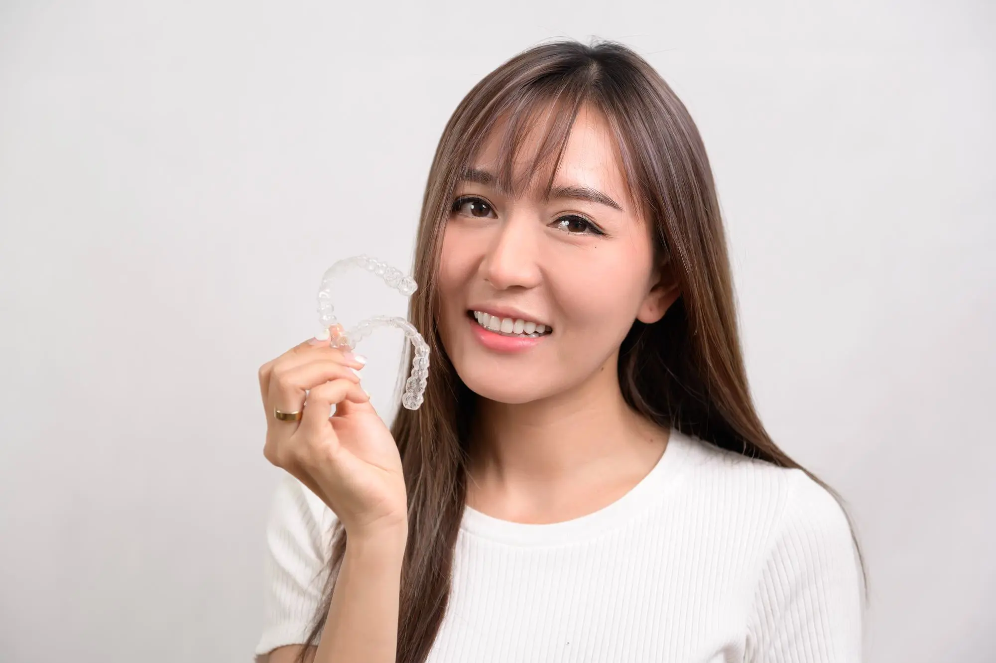 Smiling young woman with long brown hair and bangs wearing a white sweater, holding a clear orthodontic aligner and displaying a confident smile against a light background, representing successful orthodontic treatment outcomes with insurance coverage at Sweet Smiles Orthodontics in Arlington, MA
