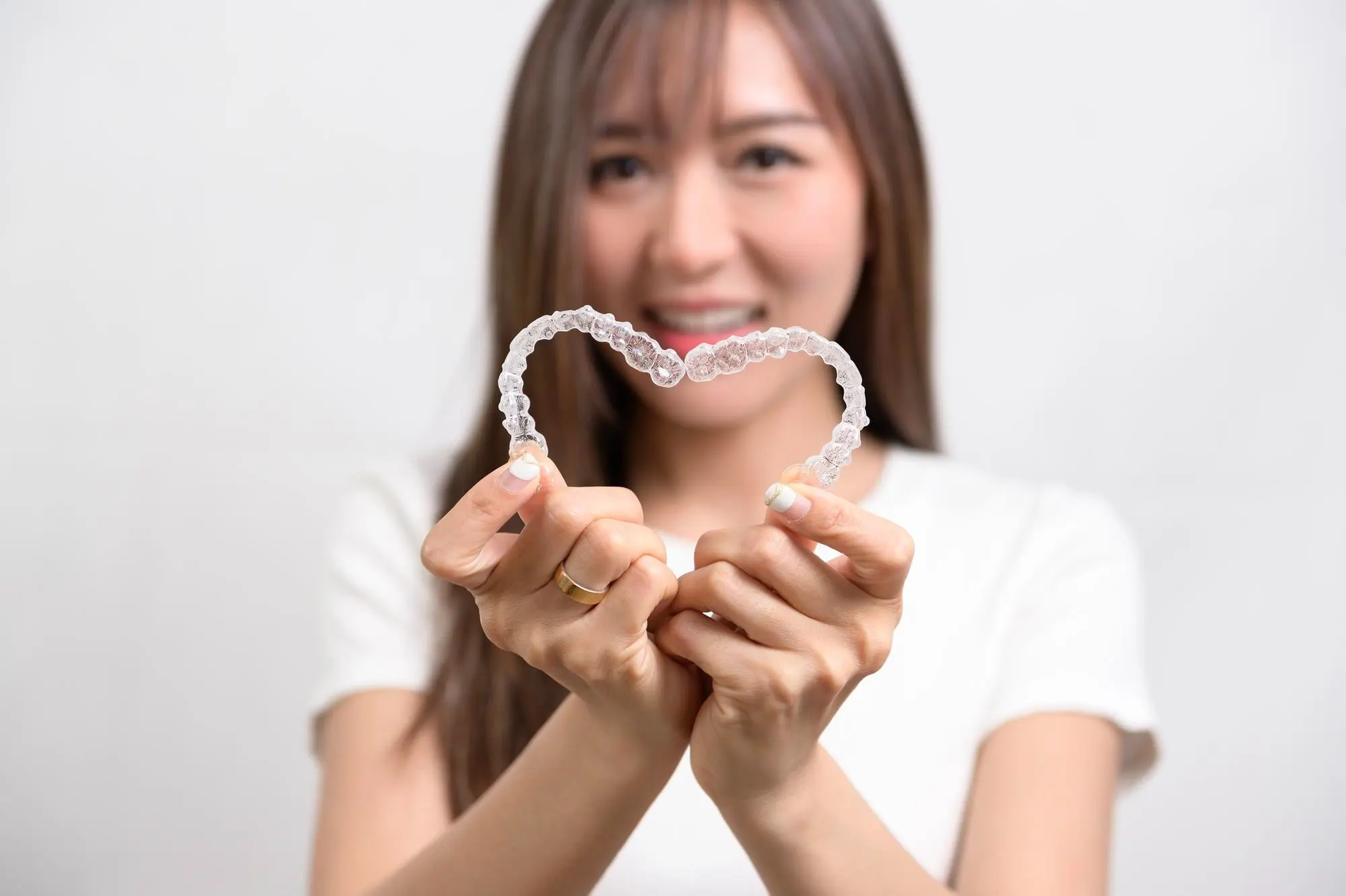 Smiling woman in white forms a heart with aligners, highlighting Invisalign vs Braces at Sweet Smiles Orthodontics in Arlington, MA.