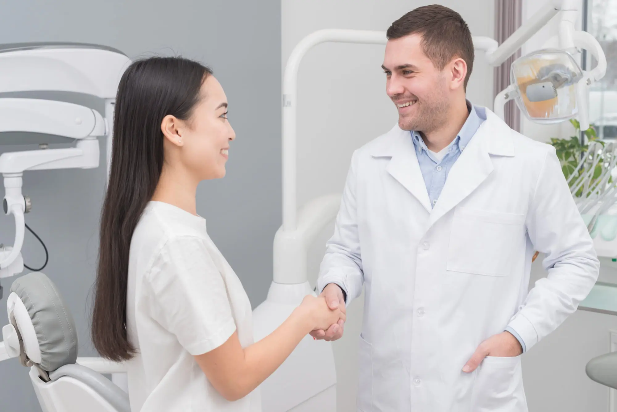A female patient in a white top shaking hands with a male orthodontist in a white lab coat in a modern orthodontic office, with dental equipment and treatment chairs visible in the background represent orthodontic insurance plans at Sweet Smiles Orthodontics in Arlington, MA