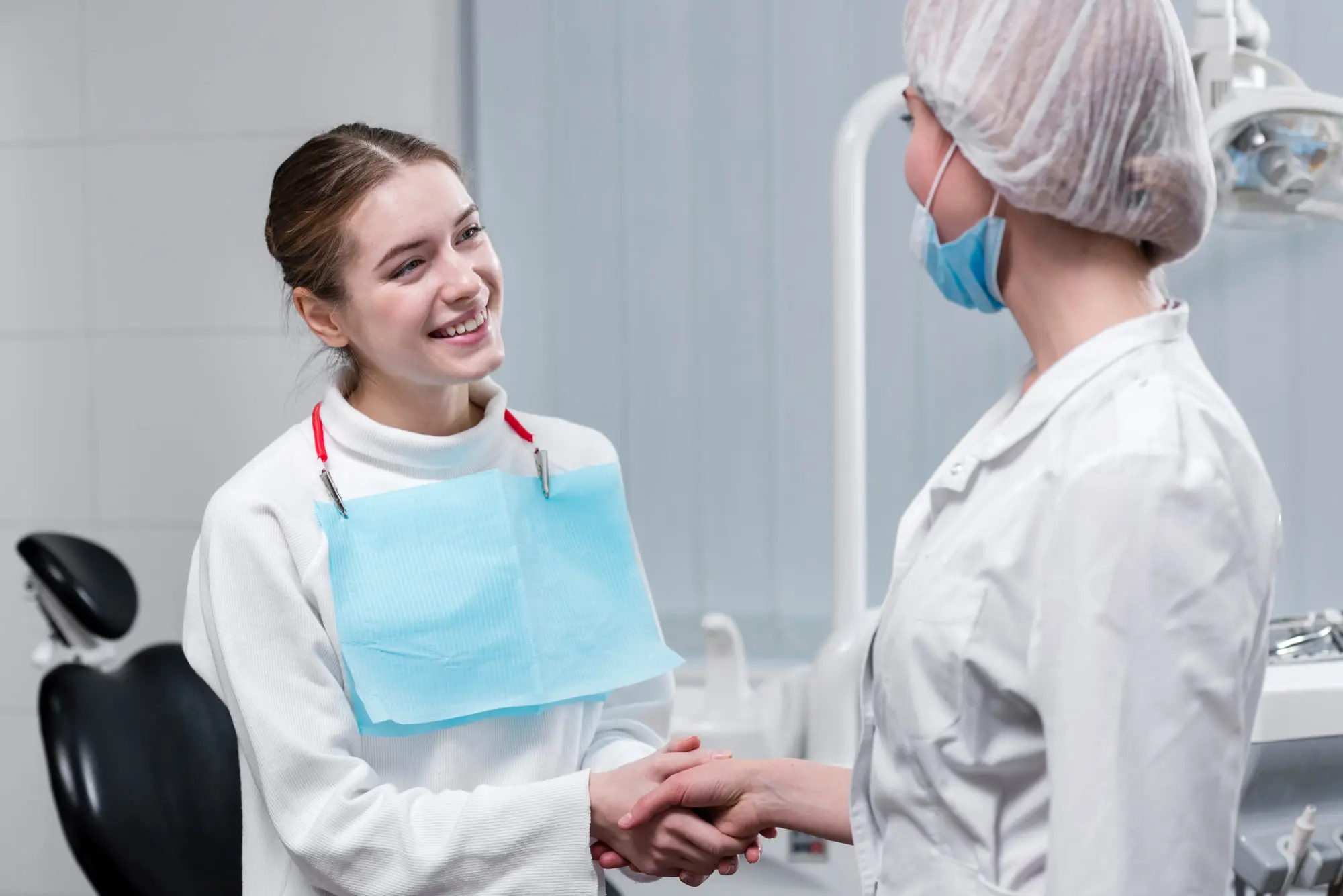 At Sweet Smiles Orthodontics in Arlington, MA, a young woman after dental care with metal braces shakes hands with a masked female dentist in a clinic.