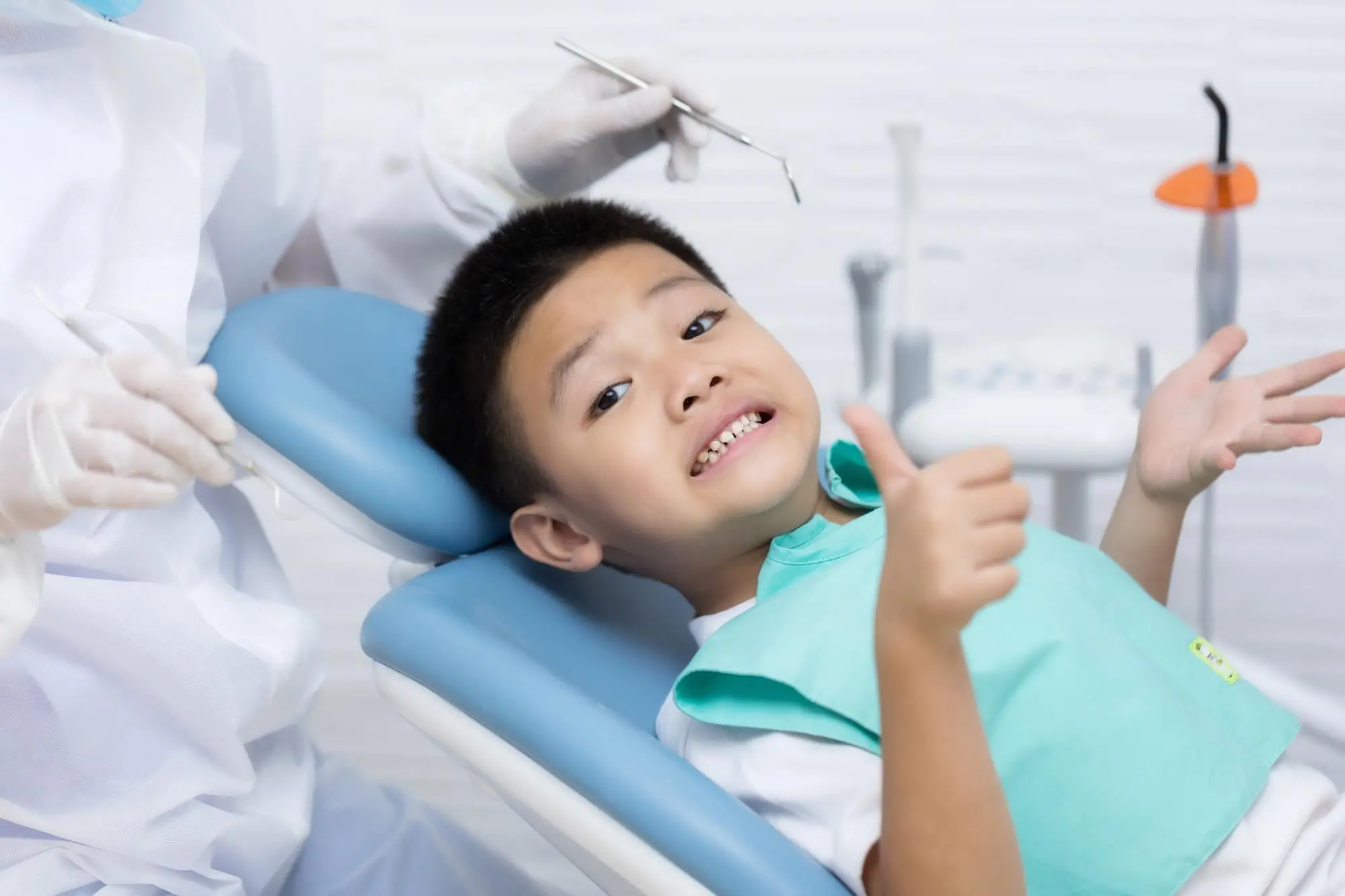 Happy young boy giving thumbs up in dental chair wearing teal bib at orthodontic office, representing positive children's orthodontic care experience at Sweet Smiles Orthodontics in Arlington, MA
