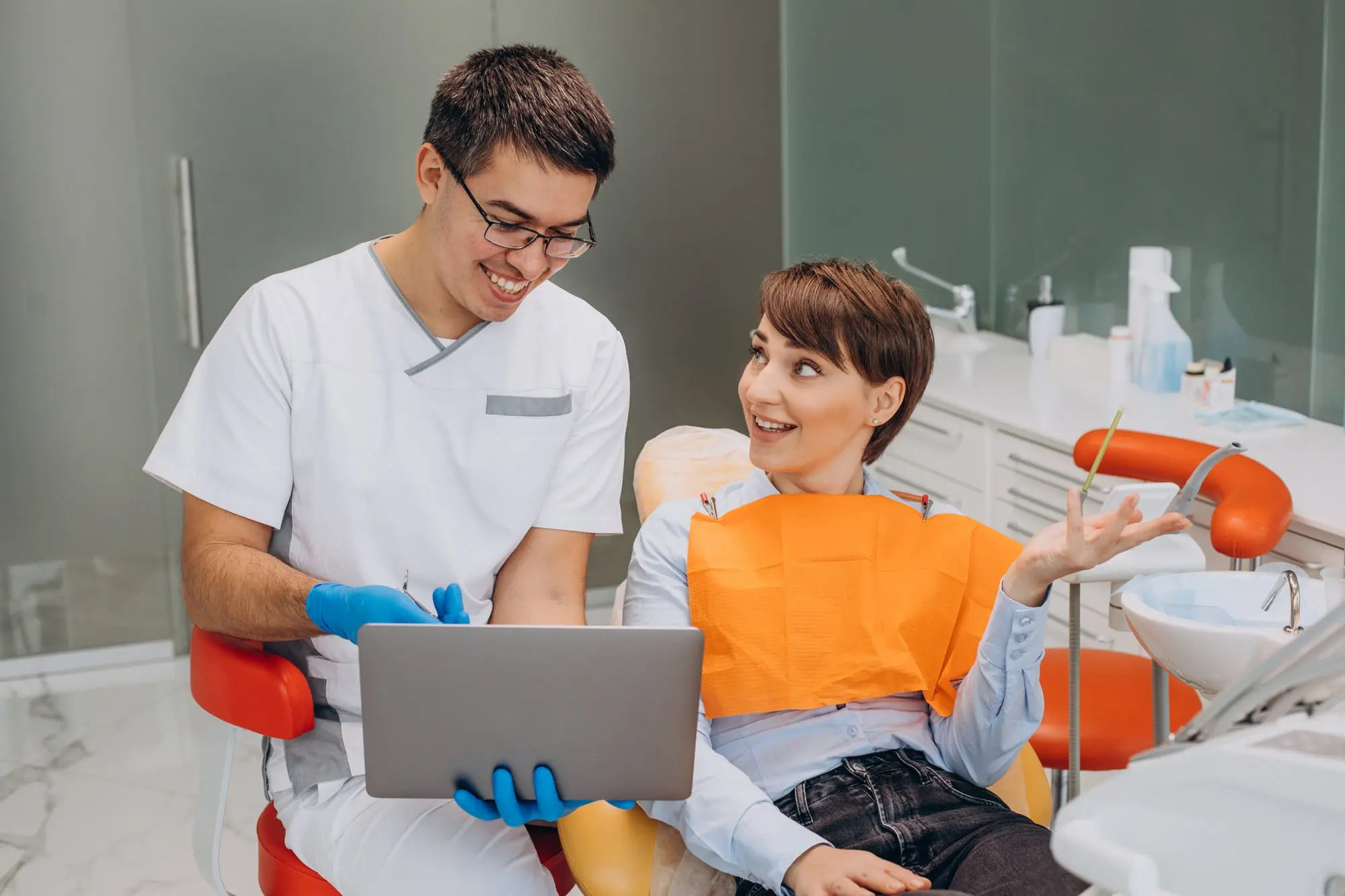 At Sweet Smiles Orthodontics in Arlington, MA, a dentist shows Invisalign options include cheap Invisalign on a laptop to a smiling patient in an orange bib.