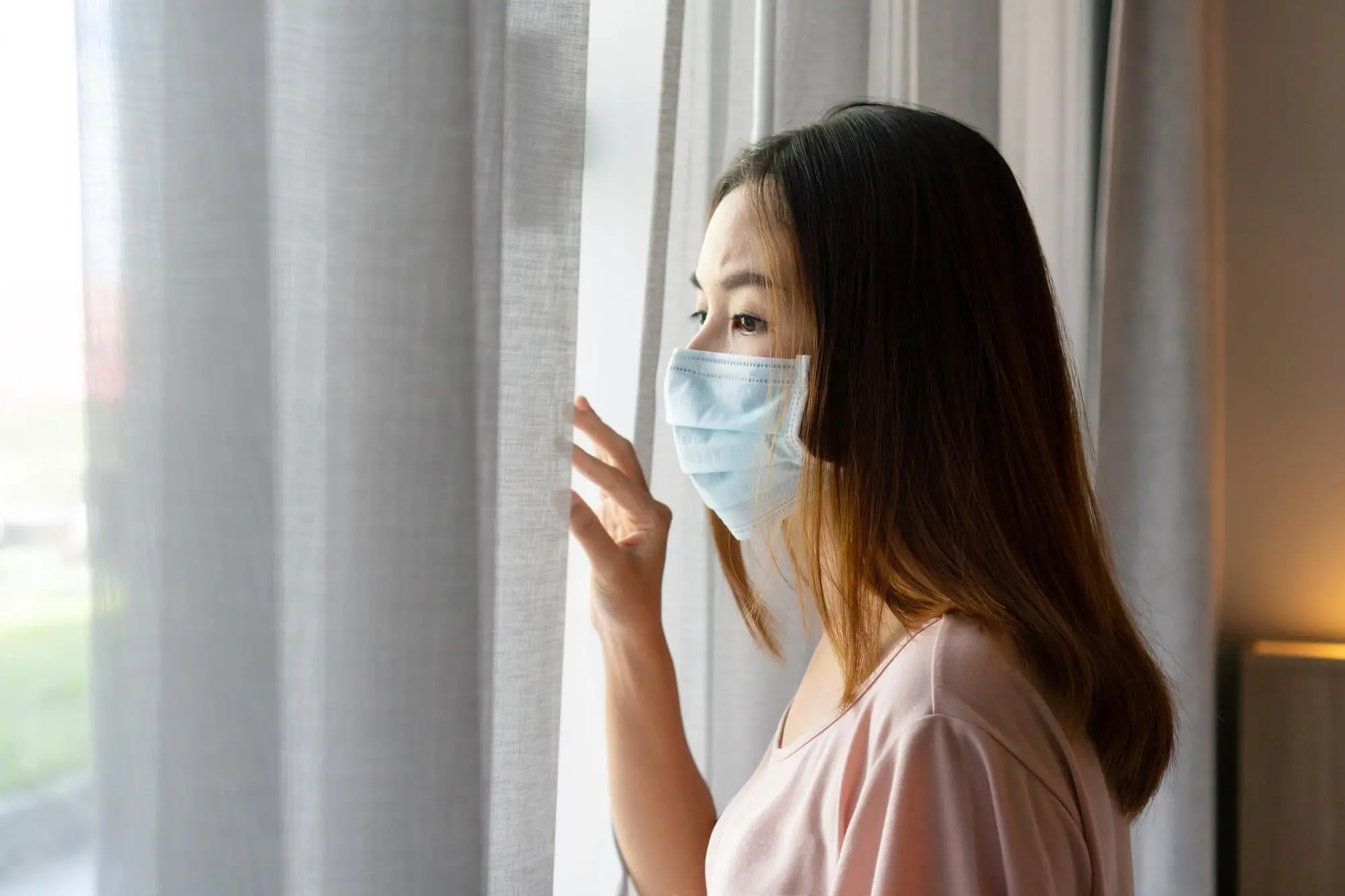 Woman in a face mask gazes out a window, gently touching the curtain, reflecting on mouth breathing at Sweet Smiles Orthodontics in Arlington, MA.