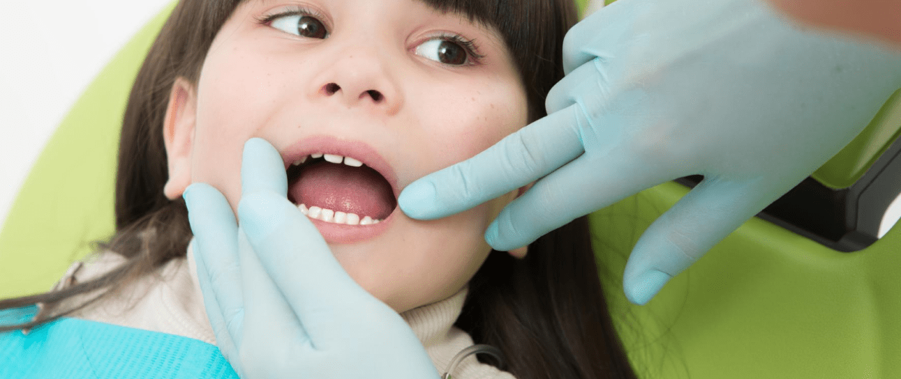 Young girl receiving orthodontic examination at Sweet Smiles Orthodontics in Arlington, MA. The child is lying in a bright green dental chair while a dental professional wearing blue latex gloves gently examines her mouth. The patient appears comfortable and relaxed during her children's orthodontic consultation