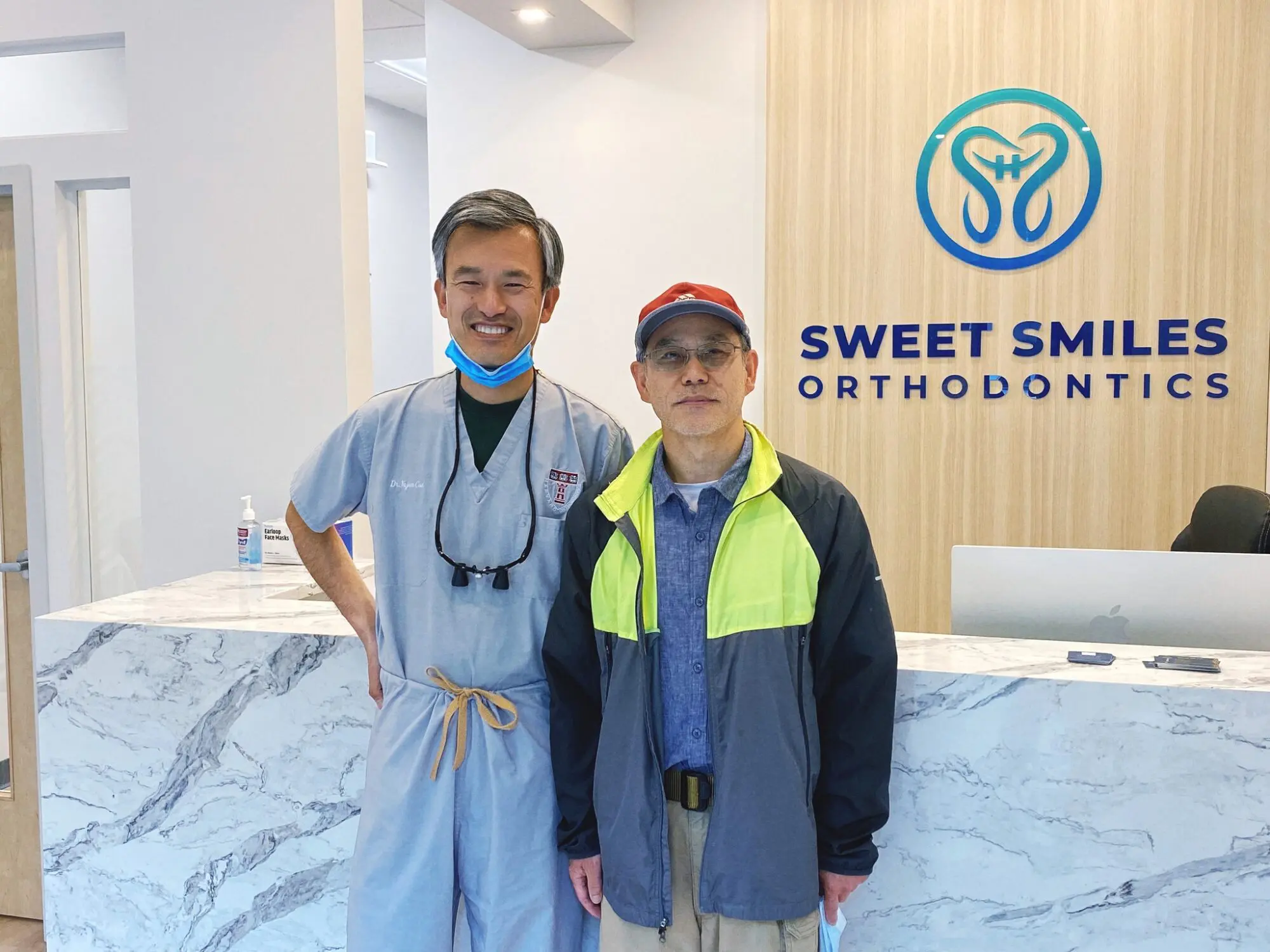 At Sweet Smiles Orthodontics in Arlington, MA, Dr. Yajun Cui in scrubs and another man in a jacket and cap smile at the desk.