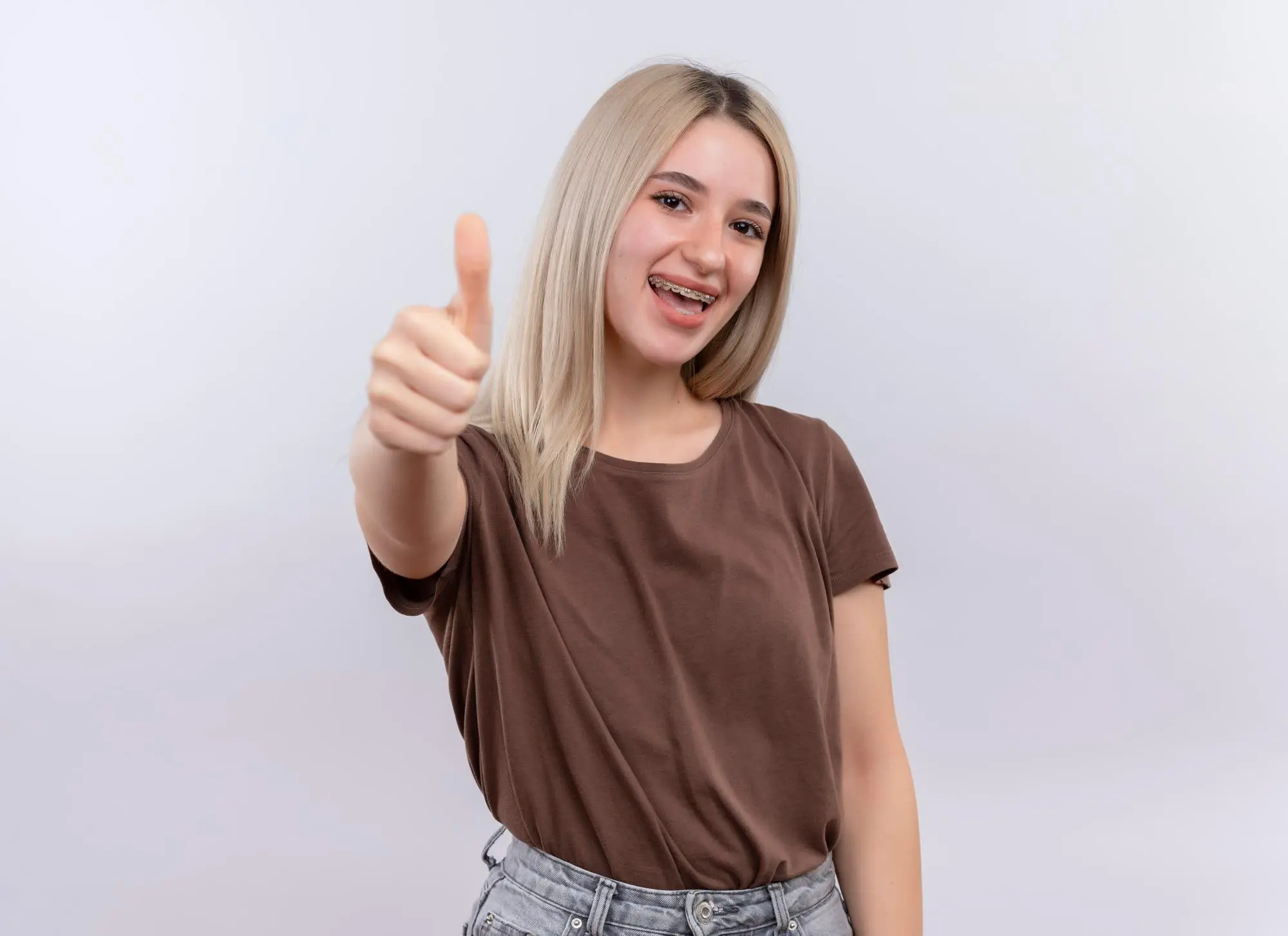 A young person with long blond hair and metal braces smiles, giving a thumbs-up for Sweet Smiles Orthodontics in Arlington, MA.