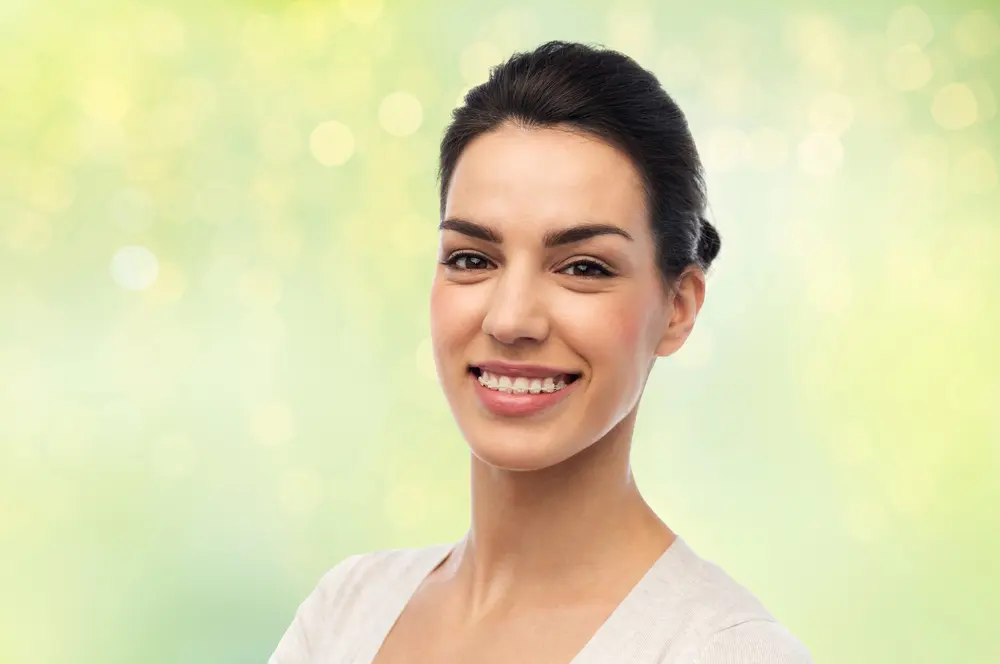 Smiling young woman with clear ceramic braces displaying her bright, confident smile against soft green background, showcasing the aesthetic appeal of tooth-colored braces available at Sweet Smiles Orthodontics in Arlington, MA