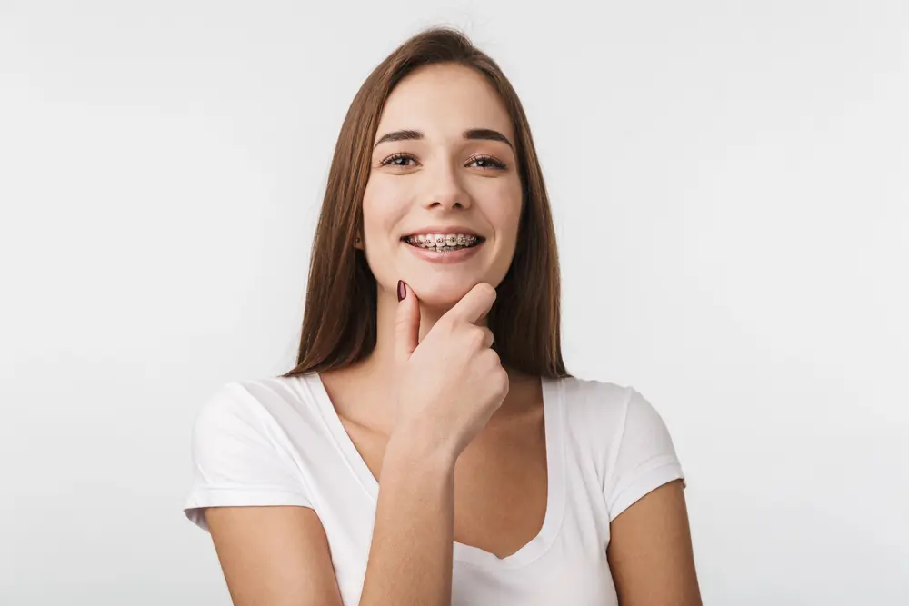Smiling young woman with long brown hair and metal braces, promoting Adult Orthodontics at Sweet Smiles Orthodontics in Arlington MA, rests her chin on her hand.