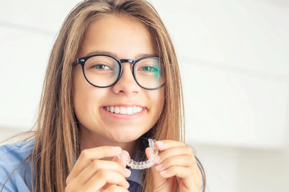 Smiling teen patient holding clear orthodontic aligner at Sweet Smiles Orthodontics in Arlington MA, showing modern invisible braces treatment for children and adolescents from Children’s Orthodontist.