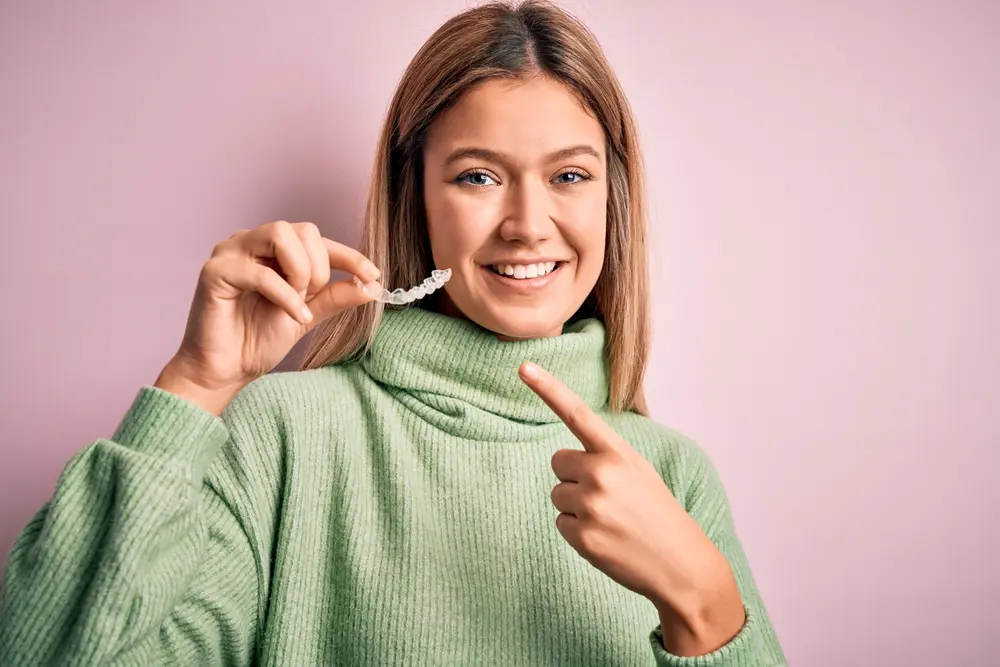 Happy young woman in green sweater giving thumbs up while holding her Invisalign clear aligner, expressing satisfaction with affordable orthodontic treatment through Invisalign Insurance benefits at Sweet Smiles Orthodontics in Arlington, MA