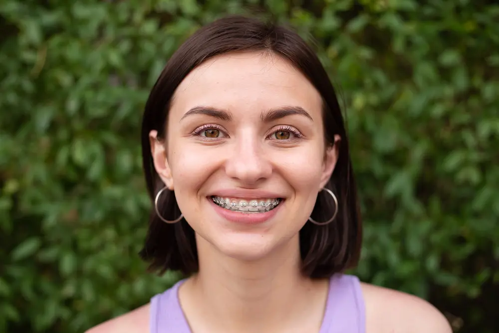 Smiling with braces, a young woman poses before greenery at Sweet Smiles Orthodontics in Arlington, MA.