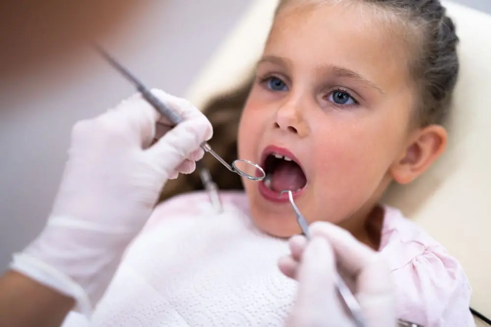 At Sweet Smiles Orthodontics in Arlington MA, a dentist examines a young girl's open mouth for tongue thrust using dental tools.