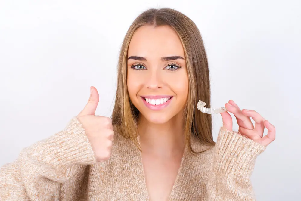 Smiling young woman gives a thumbs up and holds a clear aligner at Sweet Smiles Orthodontics in Arlington, MA.