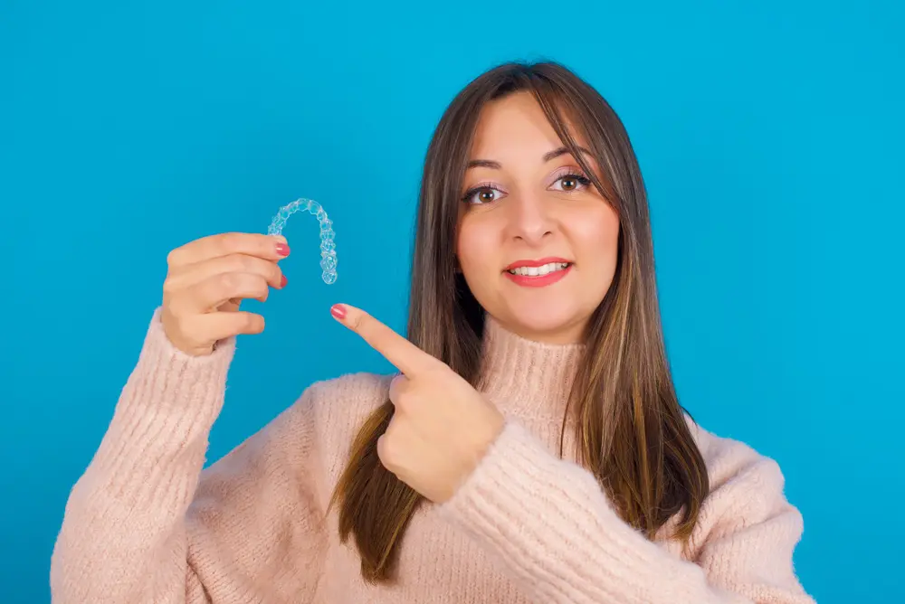 Smiling young woman in pink sweater pointing to her Invisalign clear aligner against blue background, highlighting how affordable payment plans make orthodontic treatment accessible at Sweet Smiles Orthodontics in Arlington, MA