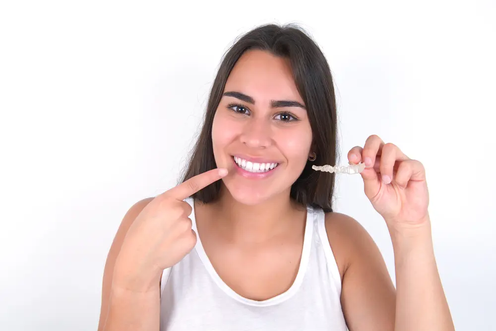 Confident young woman pointing to her perfectly straight smile while holding her Invisalign clear aligner, demonstrating successful treatment results made possible through affordable payment plan options at Sweet Smiles Orthodontics in Arlington, MA