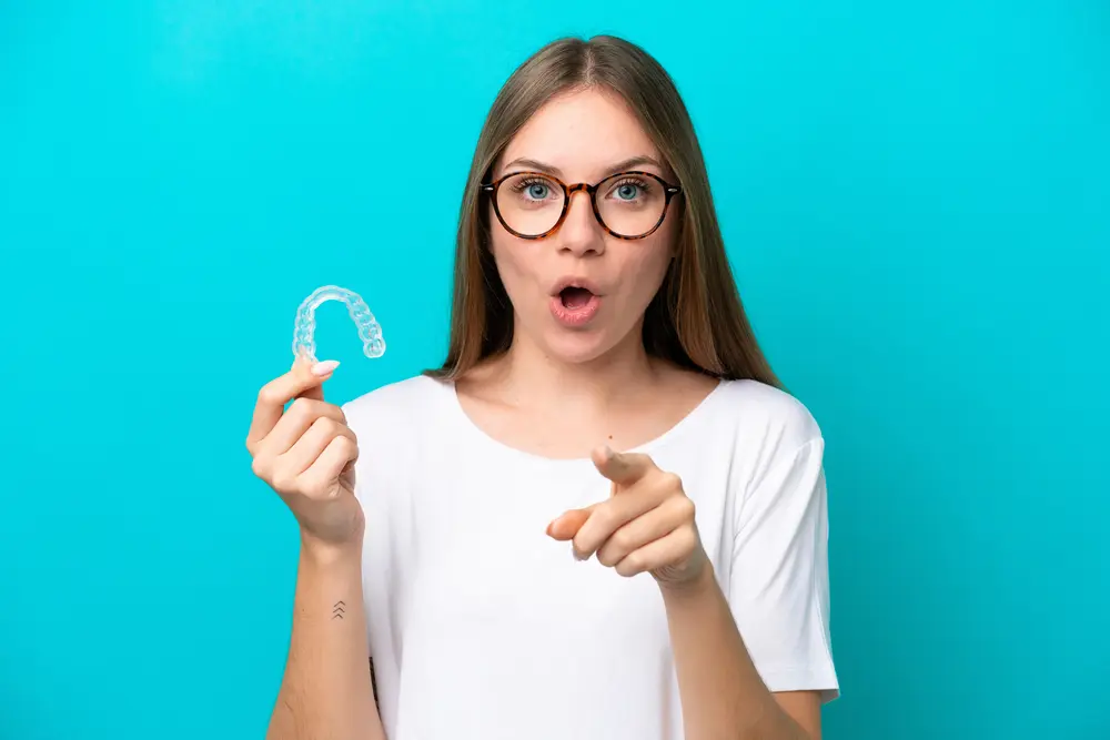 Excited young woman with glasses holding Invisalign clear aligner and pointing forward with surprised expression, representing the pleasant discovery of affordable payment plan options at Sweet Smiles Orthodontics in Arlington, MA