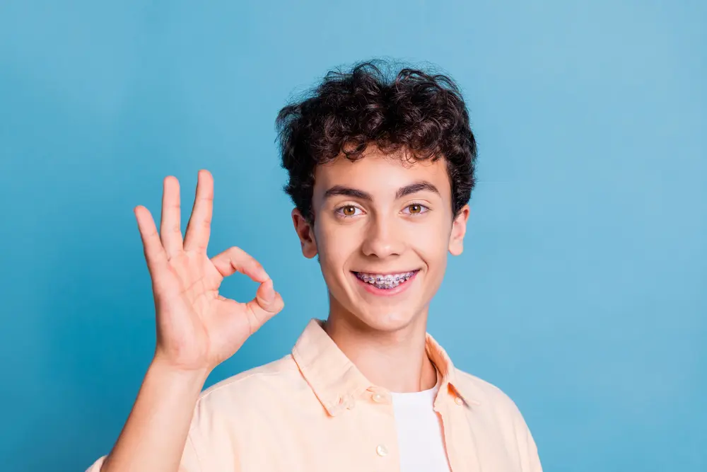Confident teenage boy with metal braces making an 'OK' hand gesture against blue background, demonstrating successful traditional braces treatment and patient satisfaction at Sweet Smiles Orthodontics in Arlington, MA