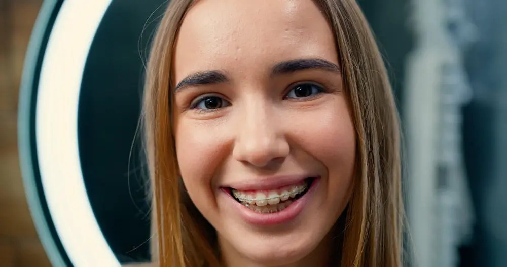 Smiling teenage girl with clear ceramic braces showing her confident smile in front of a ring light, demonstrating the discreet and aesthetically pleasing clear braces treatment available at Sweet Smiles Orthodontics in Arlington, MA