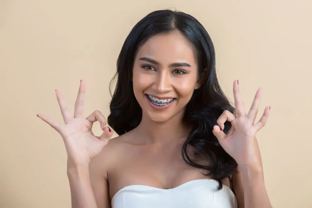 Confident young woman with colorful braces making OK hand gestures and smiling brightly, showcasing the positive attitude and satisfaction that adult patients experience with metal braces treatment at Sweet Smiles Orthodontics in Arlington, MA