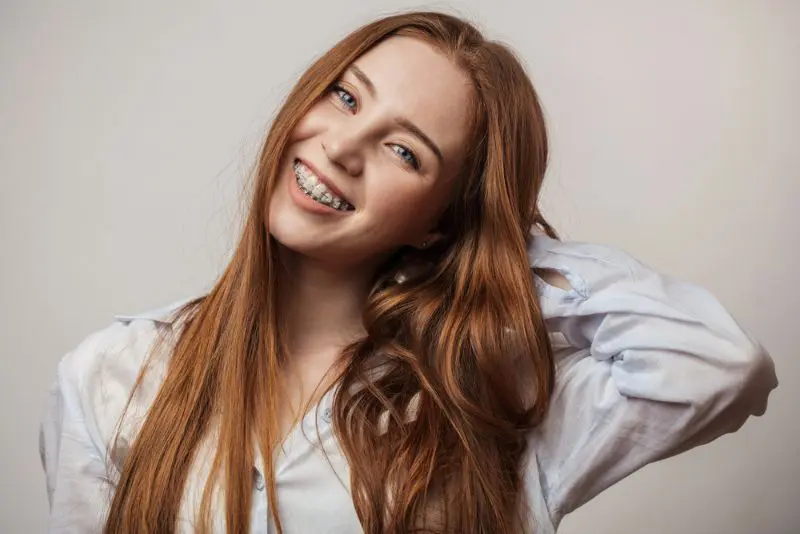 At Sweet Smiles Orthodontics in Arlington, MA, a young woman with long red hair and ceramic braces smiles in a blue shirt against a plain background.