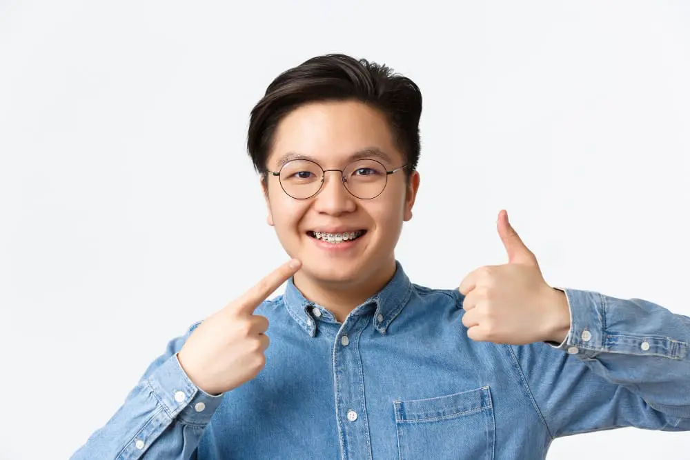 Happy young adult man wearing glasses and braces giving thumbs up while pointing to his smile, demonstrating the positive experience and confidence that comes with adult braces treatment at Sweet Smiles Orthodontics in Arlington, MA