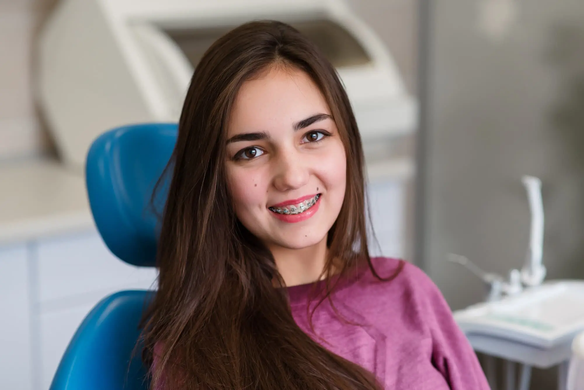 At Sweet Smiles Orthodontics in Arlington, MA, a young woman with metal braces smiles from a dental chair; equipment is visible behind her.