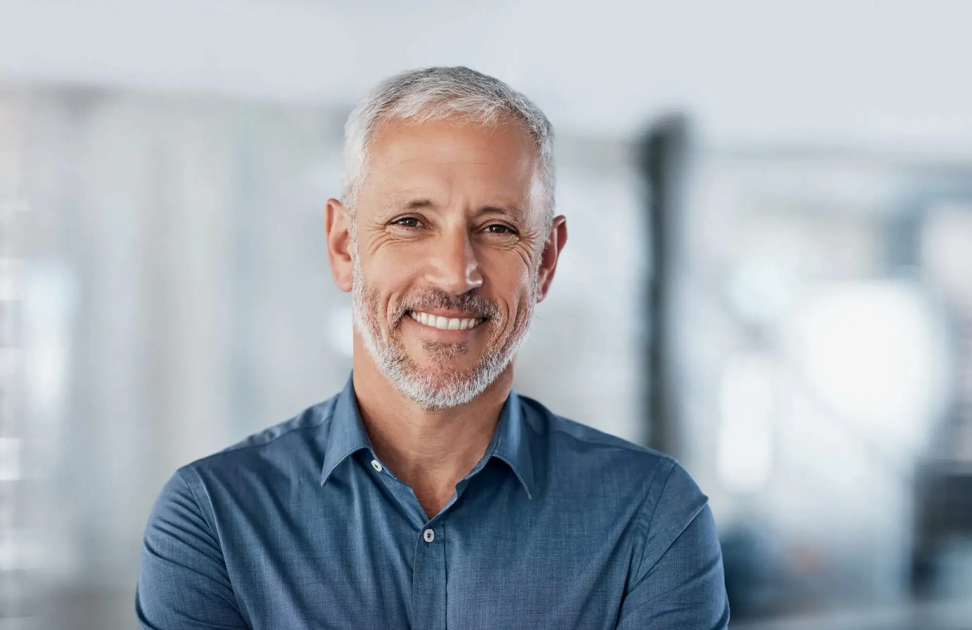 Smiling middle-aged man with gray hair, blue shirt, and showing the result from benefit adult braces at Sweet Smiles Orthodontics in Arlington, MA.