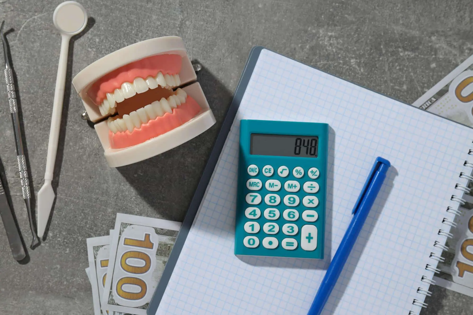Dental tools, model, calculator, pen, notebook, and $100 bills on a gray surface represent a braces payment plan at Sweet Smiles Orthodontics in Arlington, MA.