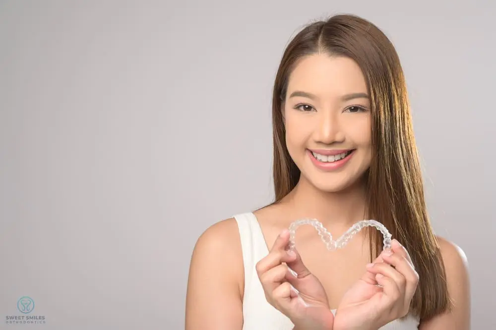 A smiling young woman holds heart-shaped clear aligners in front of her chest at Sweet Smiles Orthodontics in Arlington, MA - Can Invisalign Fix Overbite in Arlington, MA