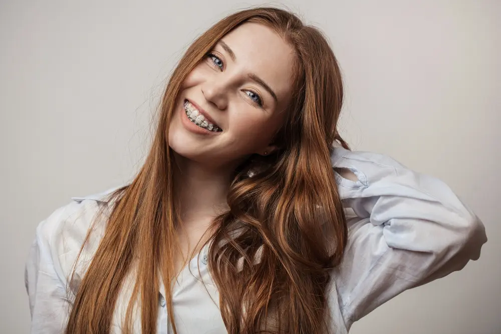 Smiling young woman with red hair, braces, and overbite at Sweet Smiles Orthodontics in Arlington, MA, holding her hair - Overbite vs. Underbite in Arlington, MA