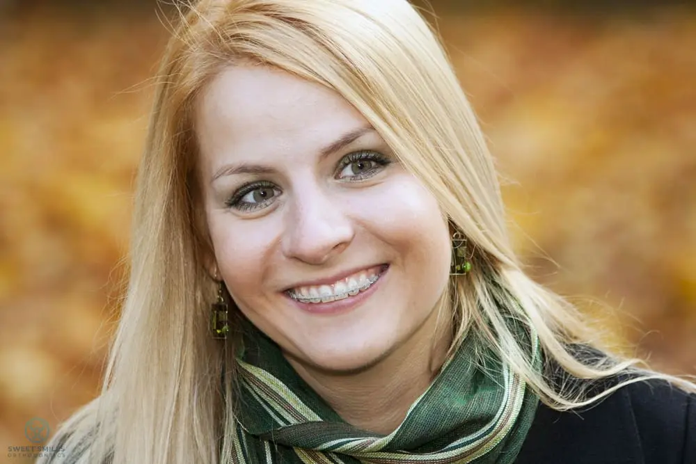 Smiling woman with blonde hair, green scarf, and braces for her overbite at Sweet Smiles Orthodontics in Arlington, MA, amid fall leaves - Overbite vs Underbite in Arlington, MA