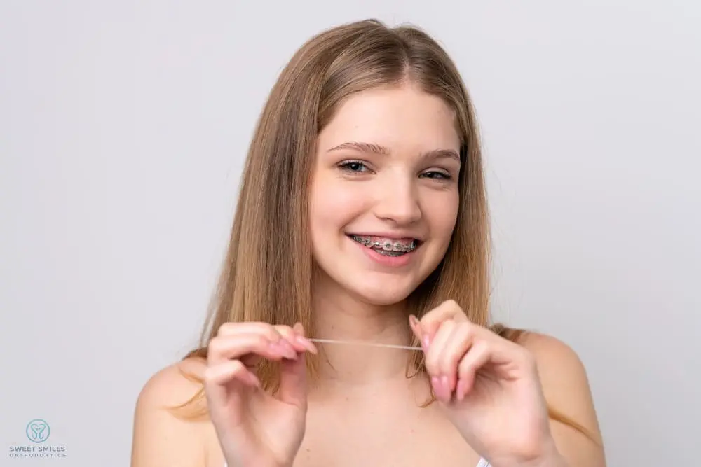 A teen girl with braces smiles, holding dental floss at Sweet Smiles Orthodontics in Arlington, MA, demonstrating braces care - How To Floss With Braces in Arlington, MA