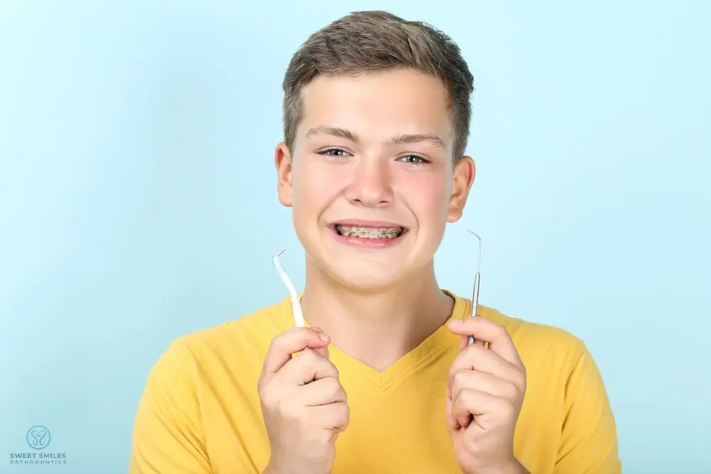 A smiling teen with braces holding dental tools promotes flossing at Sweet Smiles Orthodontics in Arlington, MA, on blue background - How To Floss With Braces in Arlington, MA