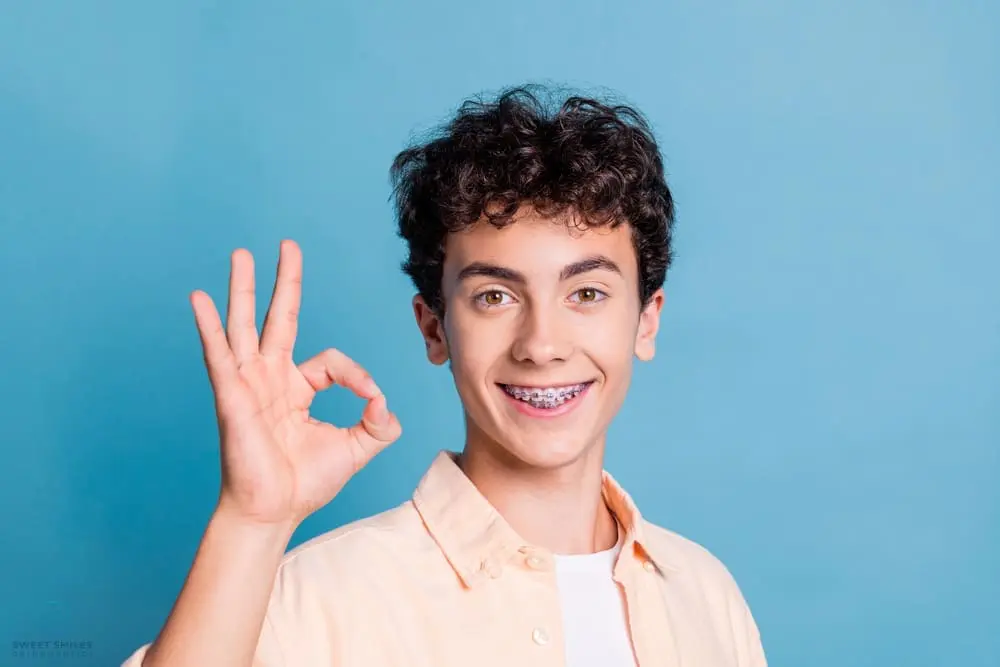 A teen with curly hair and braces gives an "OK" sign, representing Sweet Smiles Orthodontics in Arlington, MA - How To Get Braces Glue Off Teeth in Arlington, MA